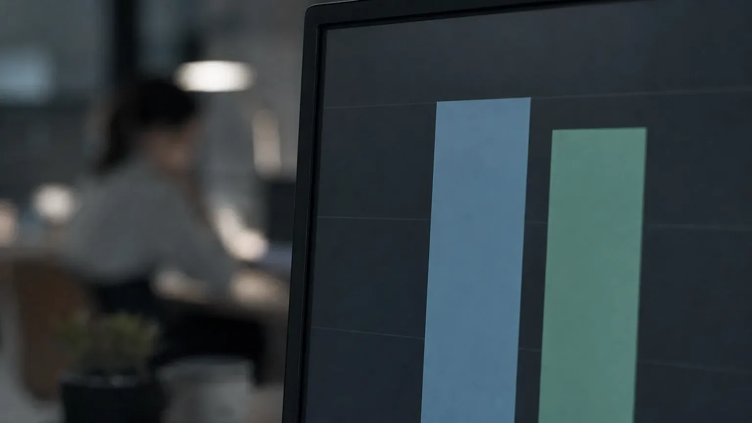 A monitor in the foreground displays a bar chart with two close-set coloured bars, while a person works at a desk under lamp light in the blurred background